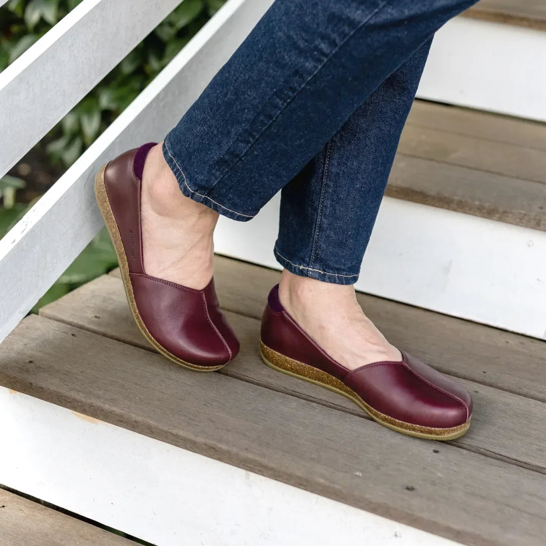 A person wearing the Women's Liesl Leather Skimmer, dark red wide toe box shoes, paired with blue jeans, stands on wooden stairs.