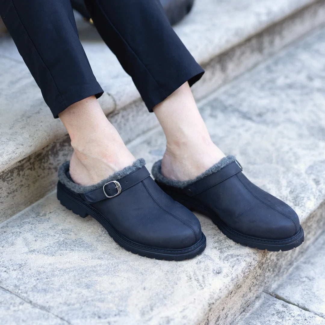 A person sits on stone steps with ankles exposed, wearing the Zermatt Wool Shearling Clog—black slip-on winter clogs crafted with vegetable-tanned leather uppers, a faux fur lining for warmth, and an elegant buckle for a refined yet cozy look.