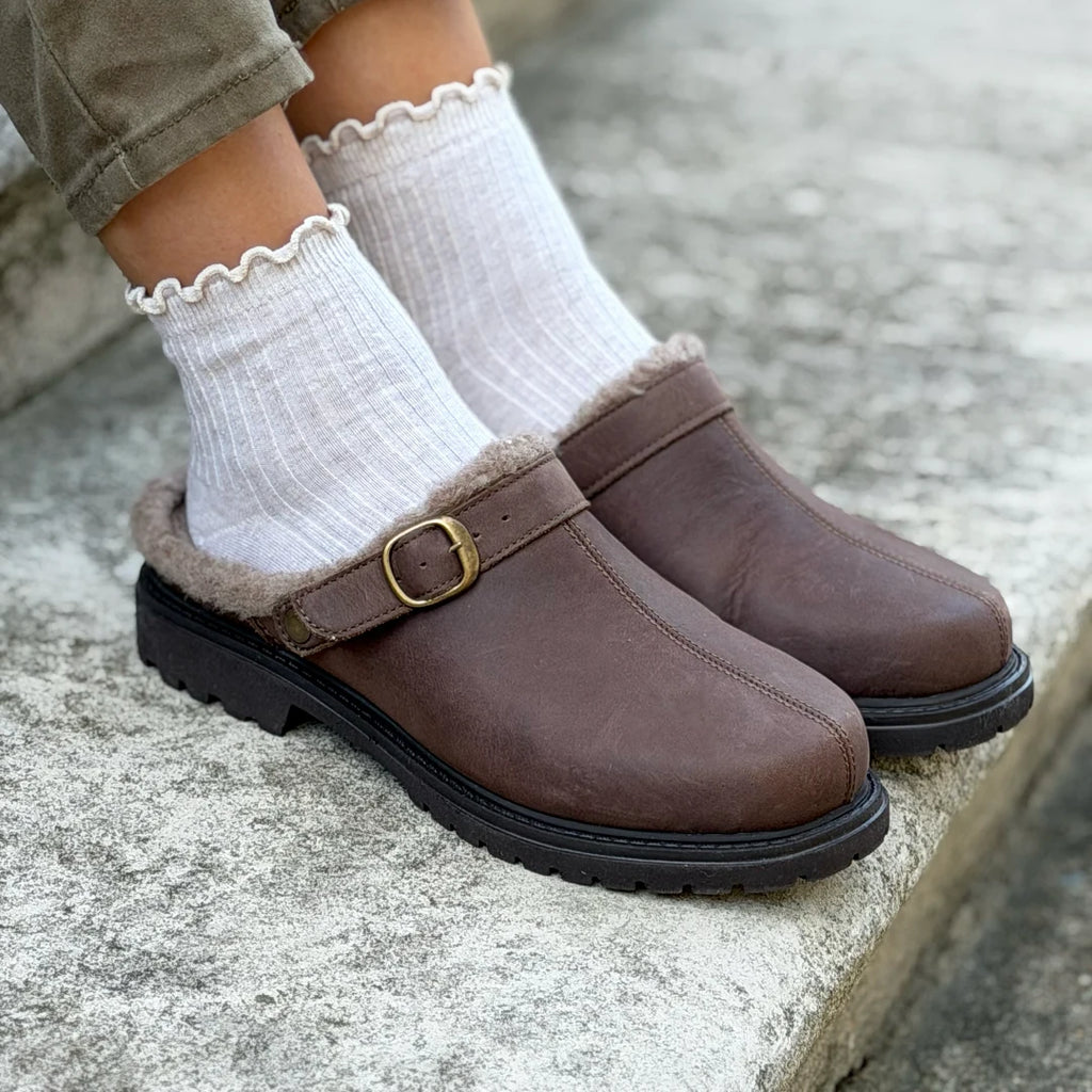 A person wearing white ruffled socks and brown Zermatt Wool Shearling Clogs, crafted from vegetable-tanned leather with buckles, sits on concrete steps.