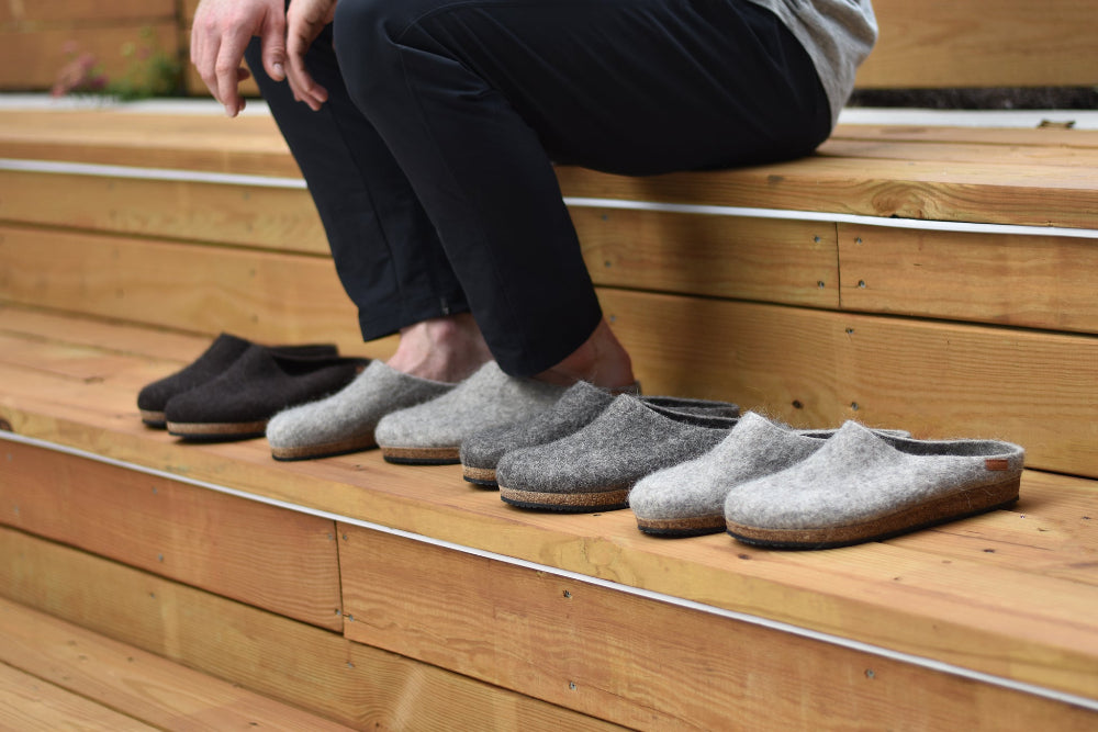 A person sits on wooden steps next to five pairs of Men's Eco Wool Clog, crafted from sustainable wool in assorted grey and brown shades.