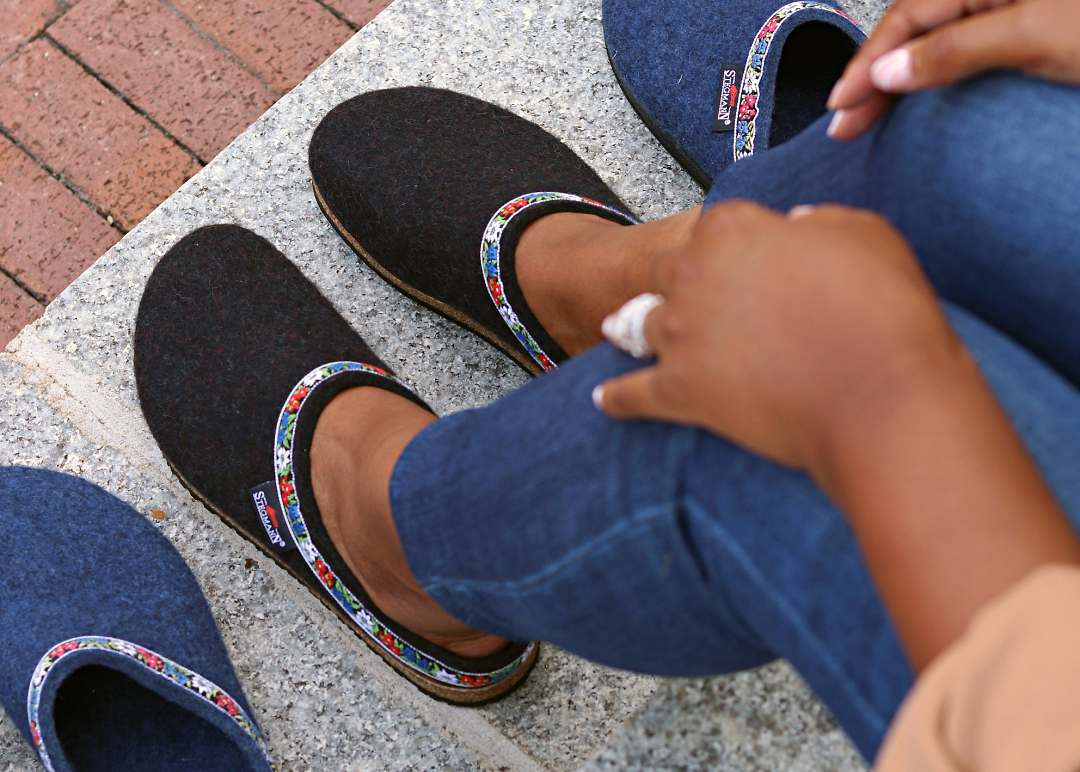 A person in black slip-on shoes with an embroidered band sits on a stone surface, while a pair of Women's Edelweiss Wool Clogs - Medium, in blue, is nearby.