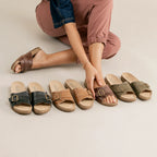 A woman sits on the floor with one hand on her ankle, next to seven pairs of Women's Ella Slide Sandal in assorted neutral tones—each pair has a contoured cork footbed and is neatly lined up.
