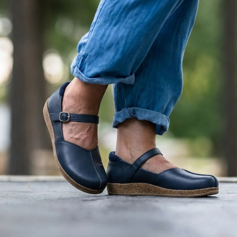 A person wearing rolled-up blue jeans and the Women's Eva Leather Mary-Jane shoes, featuring a navy blue color, wide toe box, and cork soles, walking outdoors on a paved surface.