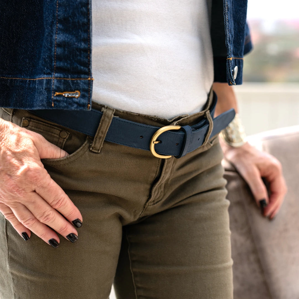 A person wearing the Women’s Matching Leather Belt, featuring a dark blue leather strap and gold buckle, styled with olive green pants, a white shirt, and a denim jacket, resting one hand on a chair.