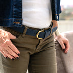 A person wearing the Women’s Matching Leather Belt, featuring a dark blue leather strap and gold buckle, styled with olive green pants, a white shirt, and a denim jacket, resting one hand on a chair.