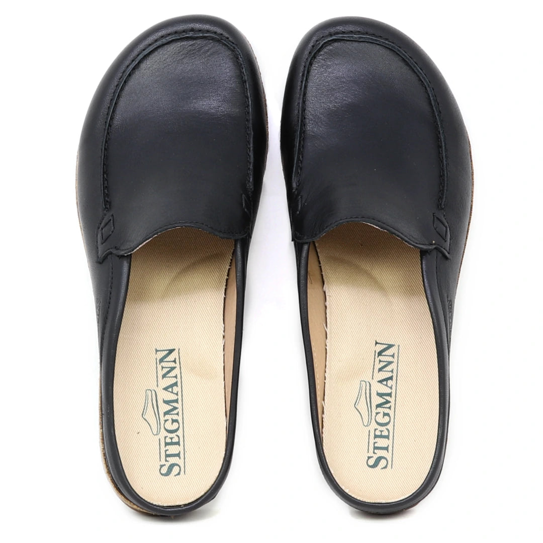 A pair of Women's Greta Leather Mule slip-on shoes in black leather, offering arch support and featuring beige insoles with the Stegmann logo and a shoe graphic.
