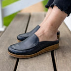 A person wearing Women's Greta Leather Mules, featuring black leather uppers and cork & latex midsoles, stands on a wooden deck with only their lower legs and feet visible.