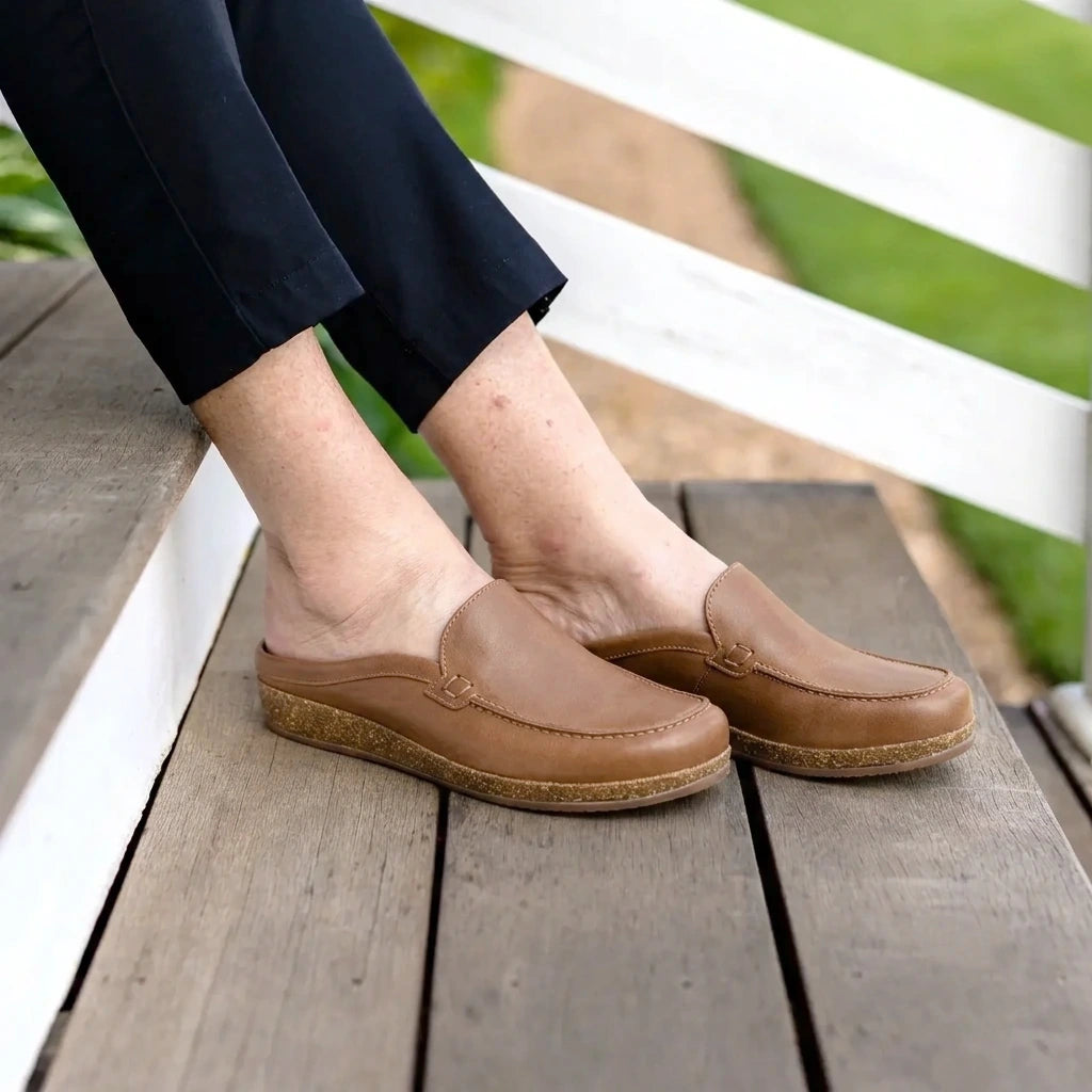 A person wearing Women's Greta Leather Mules with a cork & latex midsole sits on wooden steps outdoors, dressed in black pants.