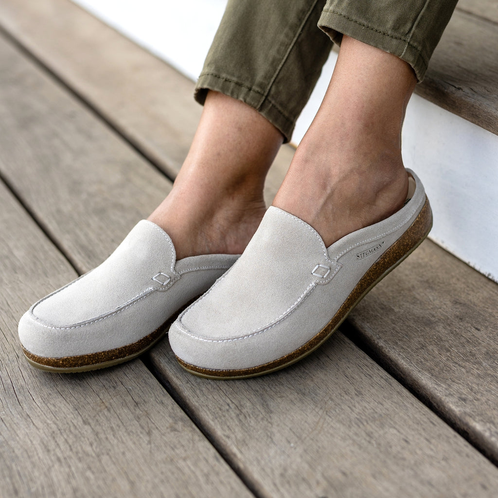 A person wearing Women's Greta Leather Mule slip-ons with cork soles and arch support, standing on wooden flooring and dressed in olive green pants.