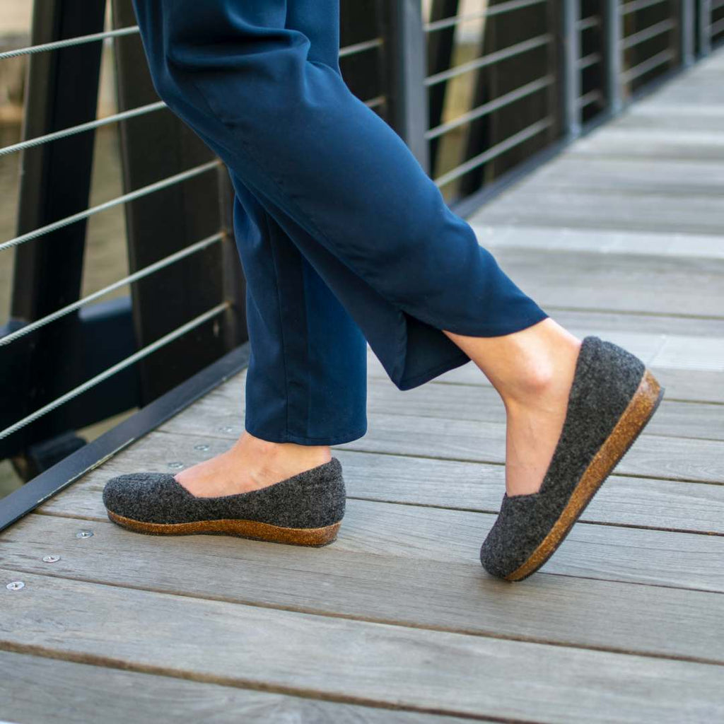 A person wearing Women's 'Liesl' Wool Skimmer - Charcoal shoes with an anatomically shaped support sole and blue pants walks on a wooden boardwalk with metal railings.