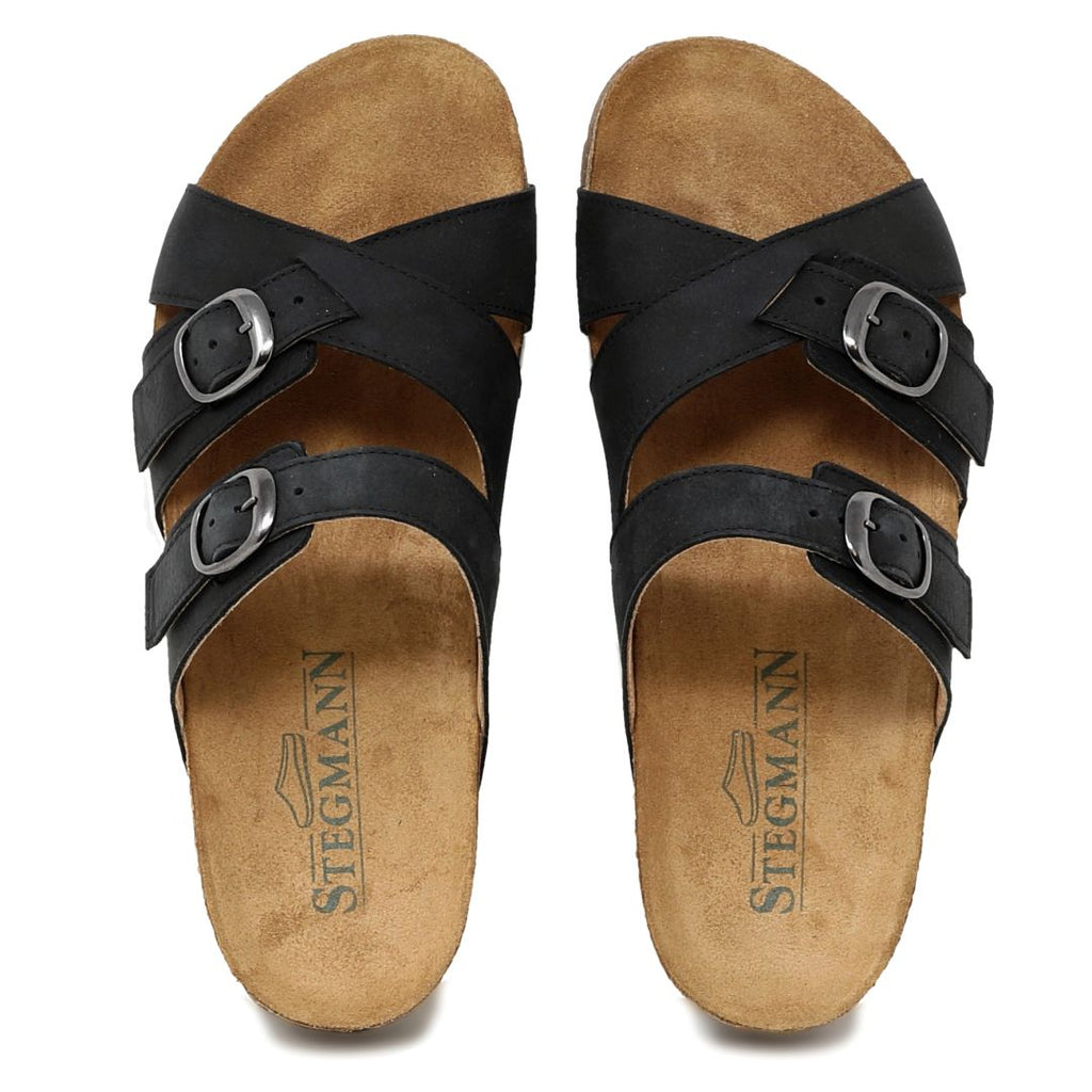 The Women's Heidi Leather Sandal features a pair of black, three-strap sandals with adjustable buckles, sustainable cork midsoles, tan cushioned soles, and an ultra-cushy footbed for enhanced comfort. The "Stegmann" branding adds elegance.