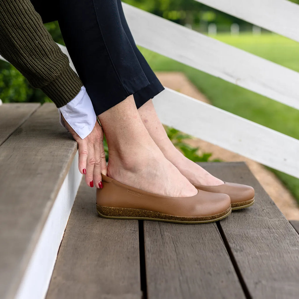 A person puts on a pair of tan Women's Helene Stretch Leather Ballet Flats while sitting on wooden outdoor steps, with green grass in the background.