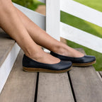A person wearing Women's Helene Stretch Leather Ballet Flats with cork soles sits on wooden steps by a white railing, green grass in the background.