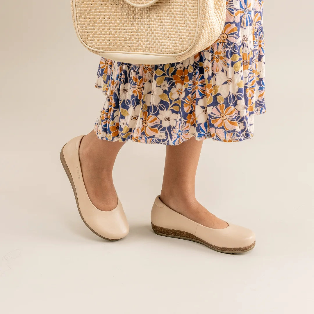 A person wearing Women's Helene Stretch Leather Ballet Flats, a floral skirt, and carrying a woven bag stands against a light background.