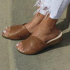A person stands on a concrete surface, wearing Women's Layna Leather Wedge Sandals, featuring a sustainable cork midsole and cushioned suede footbed, paired with white distressed jeans.