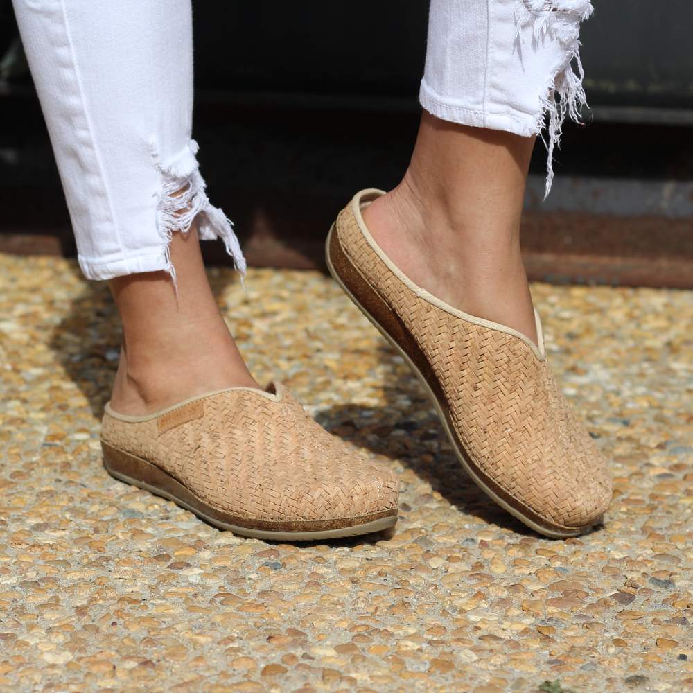 Standing on a pebble surface, someone wears the Women's Maria Woven Cork Mules in beige, featuring arch support and paired with ripped white jeans for an ideal blend of style and comfort.