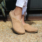 Wearing the Women's Maria Woven Cork Mule shoes with frayed white pants, a person stands on a pebble surface, enjoying the arch support of sustainable cork soles.