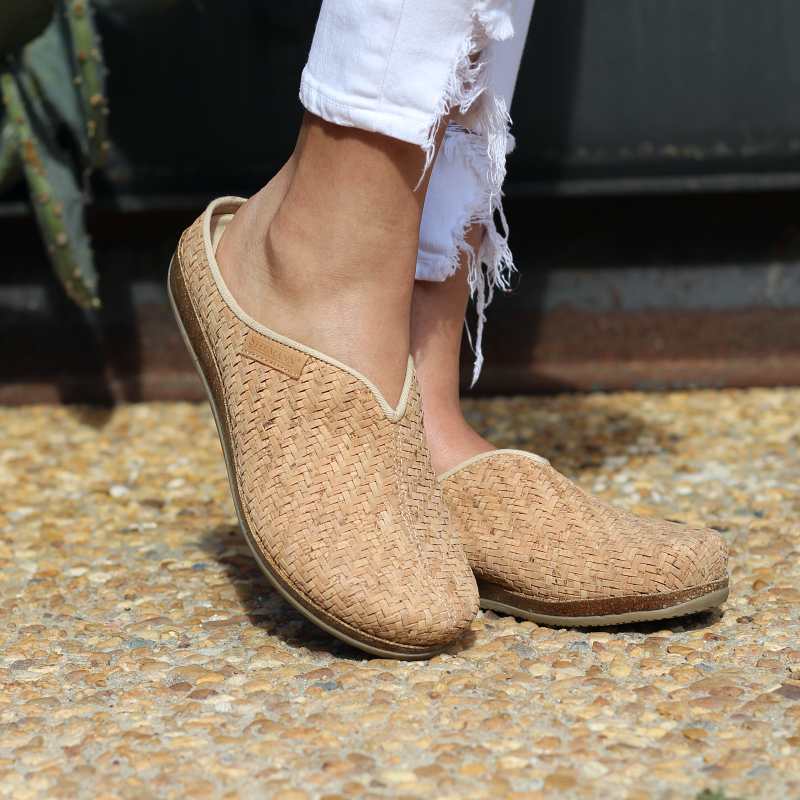 Wearing the Women's Maria Woven Cork Mule shoes with frayed white pants, a person stands on a pebble surface, enjoying the arch support of sustainable cork soles.