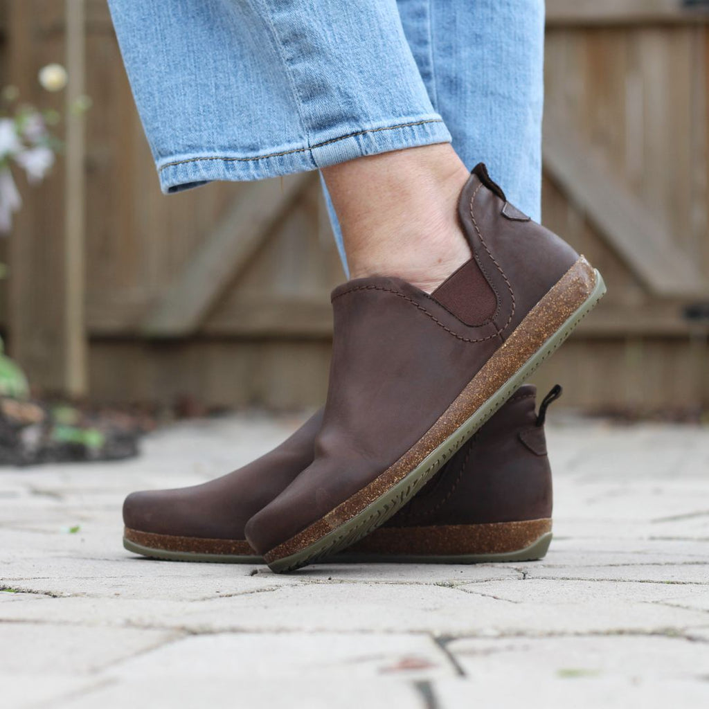 A person wears the Women's Lieben Leather Chelsea Boots on a paved surface, with a wooden fence behind them. The brown Chelsea boots made from vegetable-tanned leather perfectly complement their blue jeans.