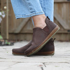 A person wears the Women's Lieben Leather Chelsea Boots on a paved surface, with a wooden fence behind them. The brown Chelsea boots made from vegetable-tanned leather perfectly complement their blue jeans.