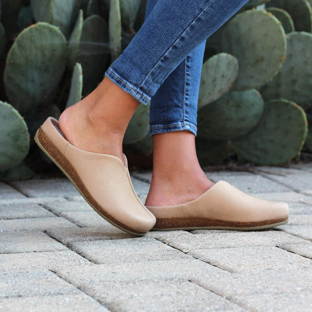 A person in the Women's Maria Leather Mule—tan with a cork & latex support sole—stands on a stone pathway, surrounded by large cactus plants.