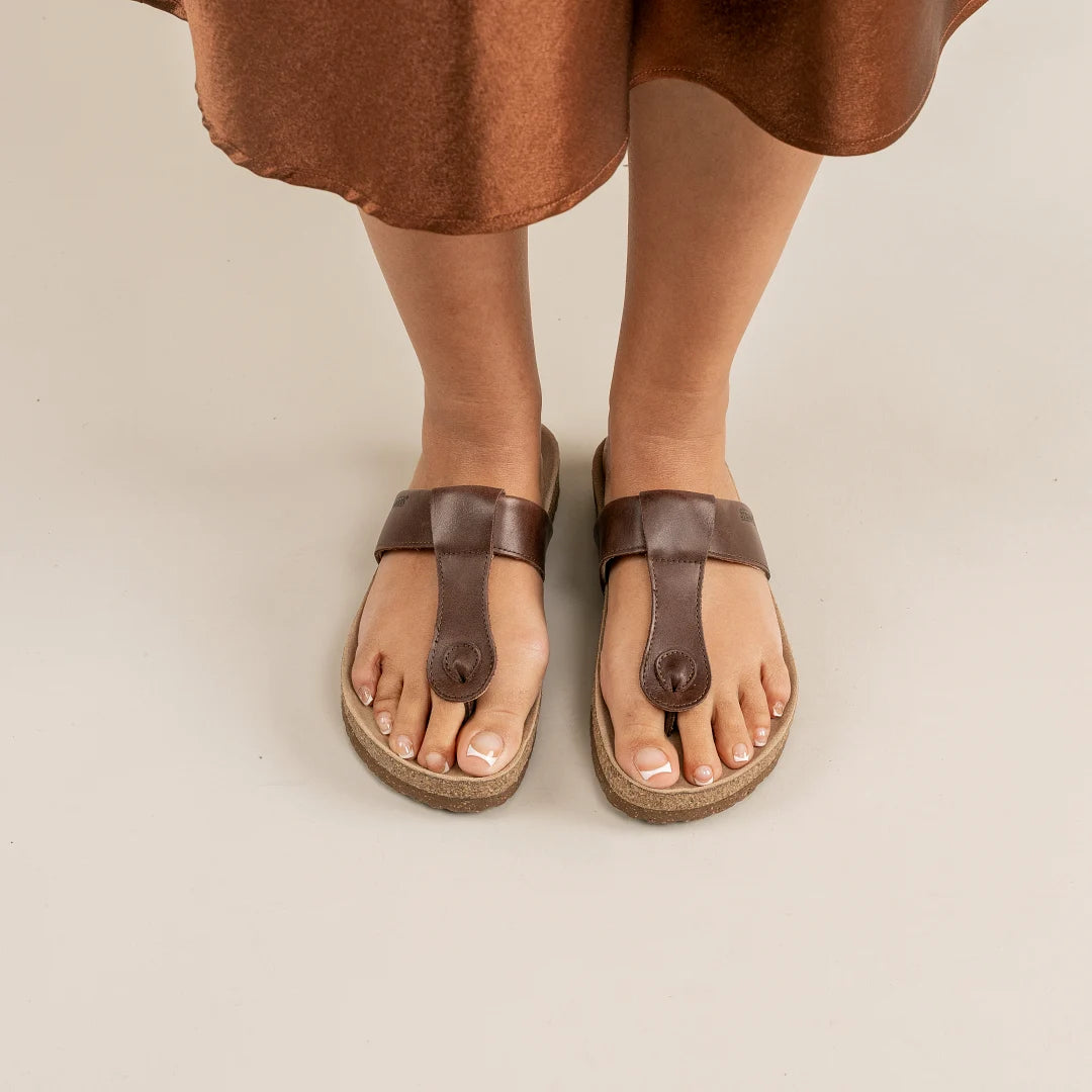 A person wearing the Women's Inga Leather Sandal, featuring a brown leather strap and padded cork footbed, stands on a light-colored surface with the hem of a brown skirt visible.