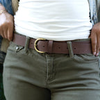 Person wearing olive green pants, a Women’s Matching Leather Belt with a gold buckle, and a white shirt, hands resting on waistband.