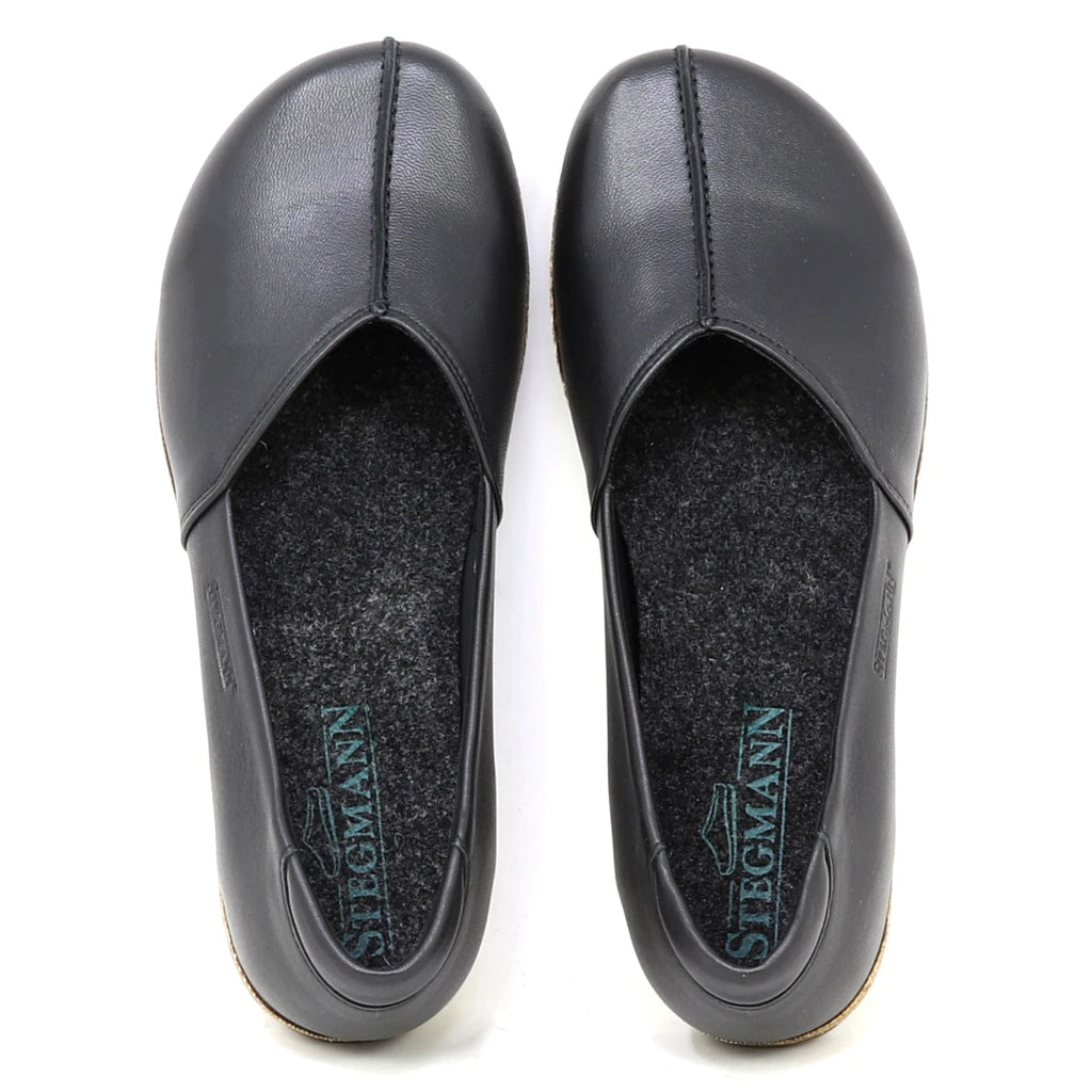 The Women's Liesl Stretch Leather Skimmer is a pair of black slip-on shoes made from buttery-soft leather, featuring a wide toe box, grey felt insoles, and "Stegmann" branding inside each shoe.