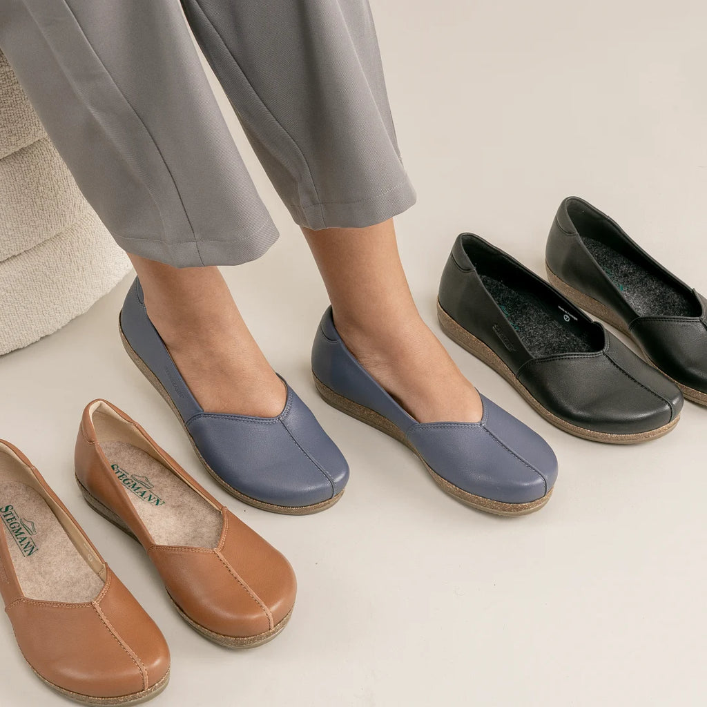A person wearing the Women's Liesl Stretch Leather Skimmer in blue, featuring a wide toe box and low wedge soles for all-day comfort, with brown and black pairs of the same style next to them on a light floor. The person is also wearing light gray pants.