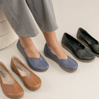 A person wearing the Women's Liesl Stretch Leather Skimmer in blue, featuring a wide toe box and low wedge soles for all-day comfort, with brown and black pairs of the same style next to them on a light floor. The person is also wearing light gray pants.