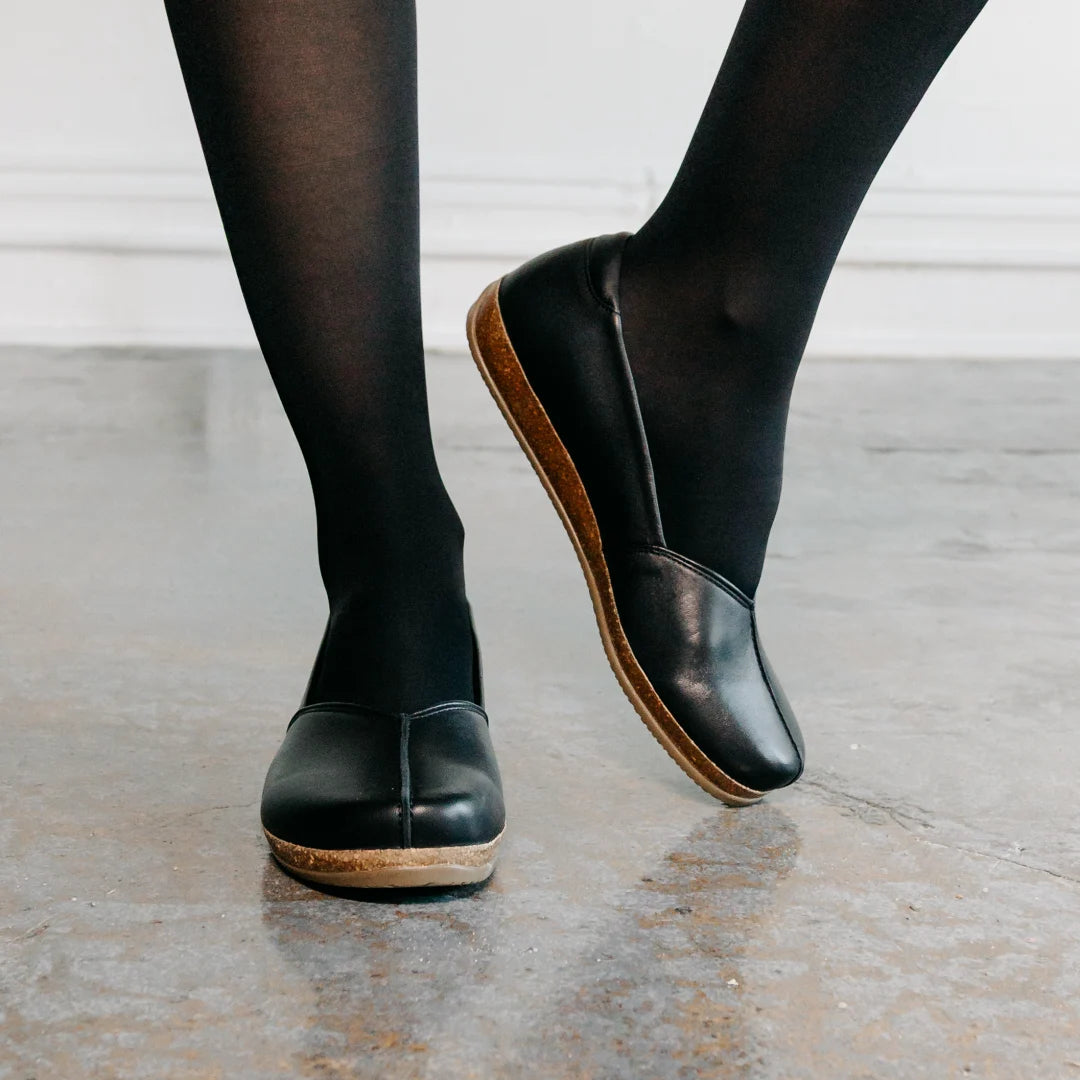 A person wears the Women's Liesl Stretch Leather Skimmer, featuring a wide toe box, paired with black tights while standing on a concrete floor with one foot slightly raised and crossed in front of the other.