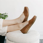 A person wears the Women's Liesl Stretch Leather Skimmer—tan slip-on shoes featuring a wide toe box and cork soles—while resting their feet on a textured white ottoman, with part of a green plant visible in the background.