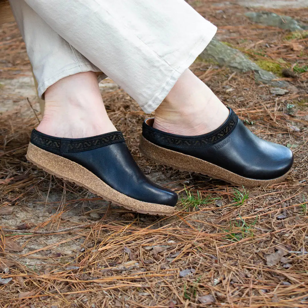 Relaxing outdoors on pine needles, a person wears the Women's Graz Leather Clog with black uppers and wedge cork soles. The vegetable-tanned leather clogs are bunion-friendly, stylishly paired with slightly rolled-up beige pants for added comfort.