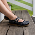 A person wearing Women's Marielle Stretch Leather Mary-Jane shoes with double straps, cork soles, and excellent arch support sits on a wooden bench outdoors.