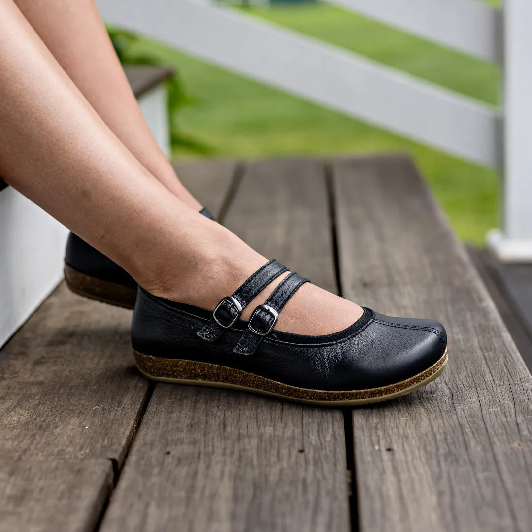 Wearing Women's Marielle Stretch Leather Mary-Jane shoes with double buckle straps, known for arch support, a person sits on a wooden deck with grass in the background.