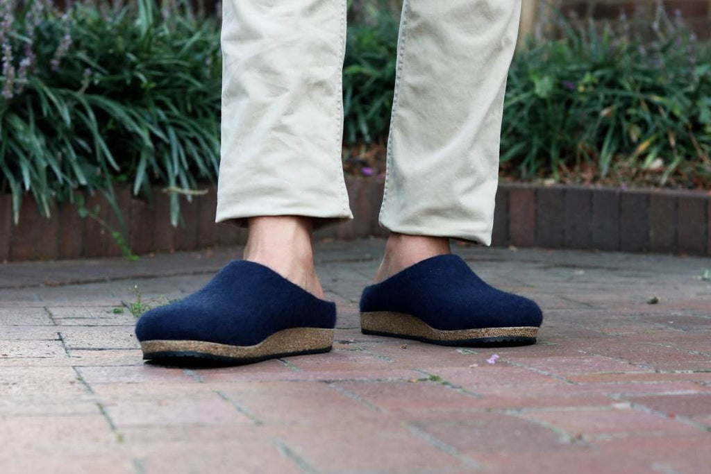 Wearing beige pants, a person in Men's Essenz Wool Clogs with a sustainable cork/latex footbed stands on a brick pathway, surrounded by lush green foliage.