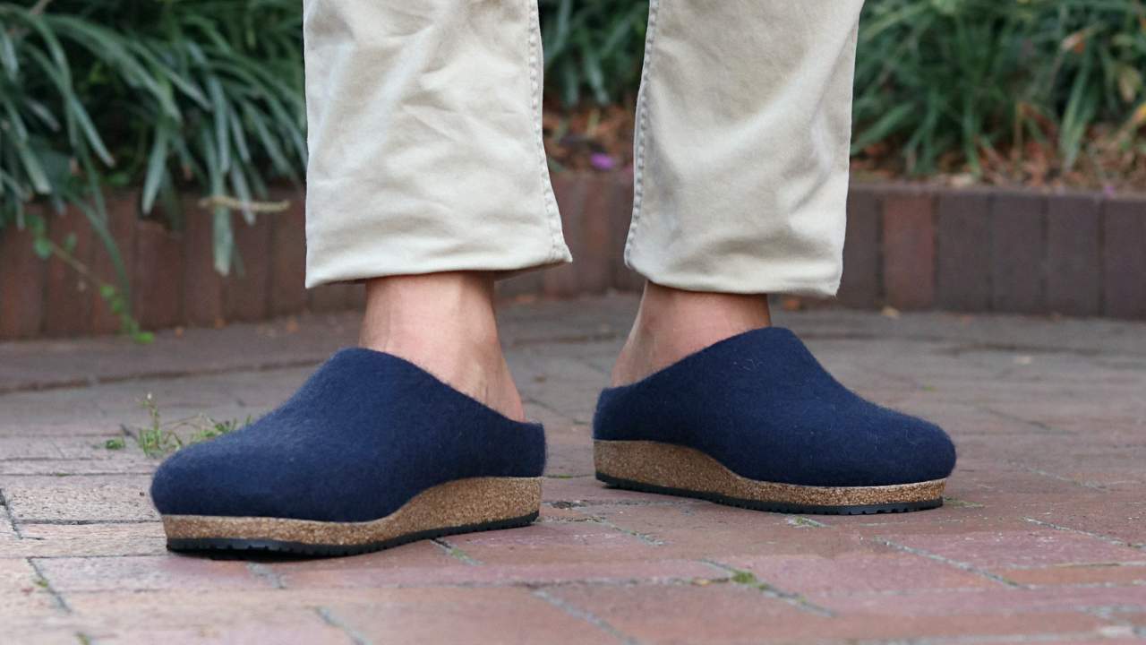 A person wearing beige pants and navy blue Men's Essenz Wool Clog shoes with cork soles and a leather tag stands on a brick walkway with greenery in the background.