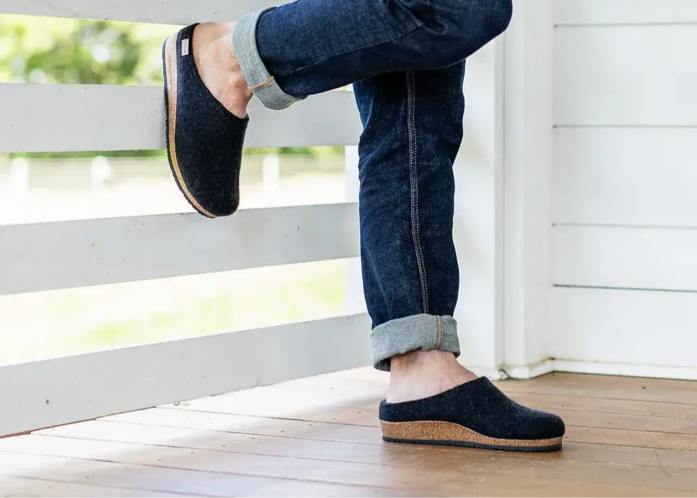 A person in dark Men's Essenz Wool Clogs with a leather tag and rolled-up jeans leans against a white wooden porch railing on a wooden floor.