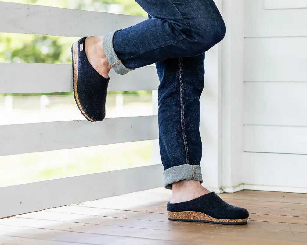 A person in dark Men's Essenz Wool Clogs with a leather tag and rolled-up jeans leans against a white wooden porch railing on a wooden floor.