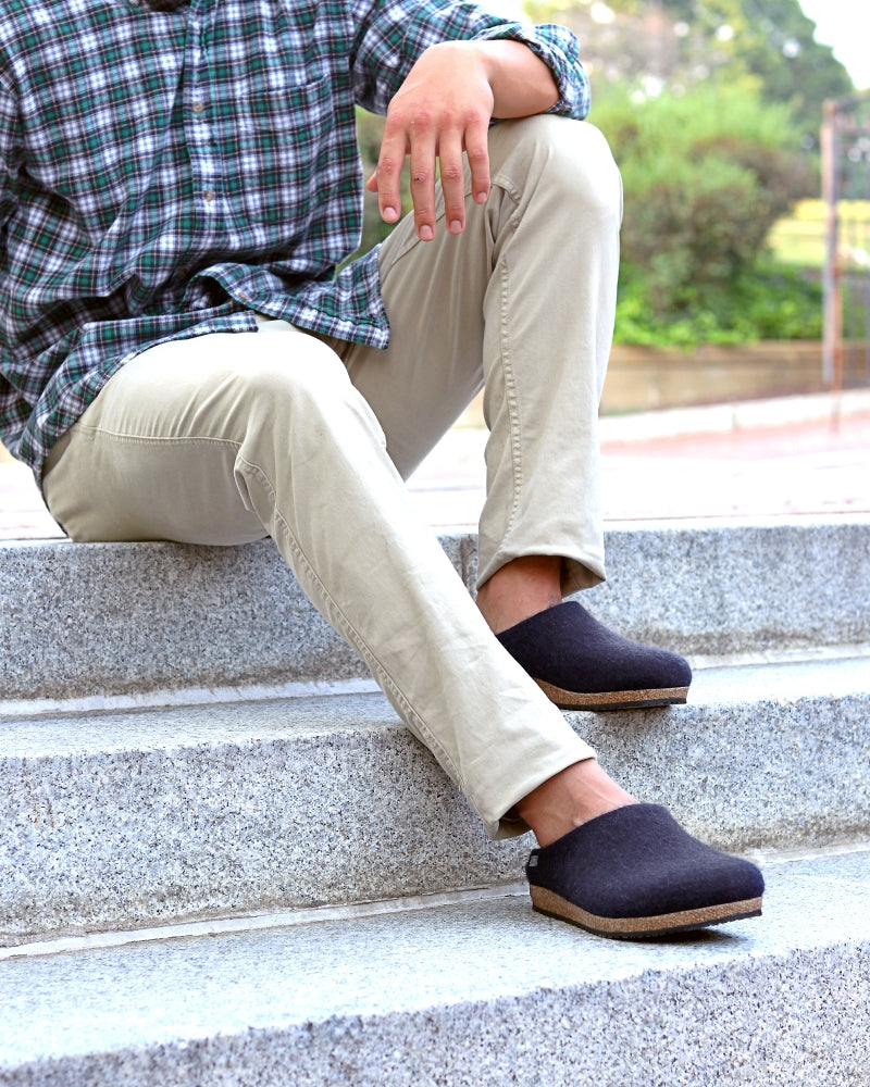 A person in a plaid shirt, beige pants, and Men's Essenz Wool Clogs sits on concrete outdoor steps, one arm resting on their knee.