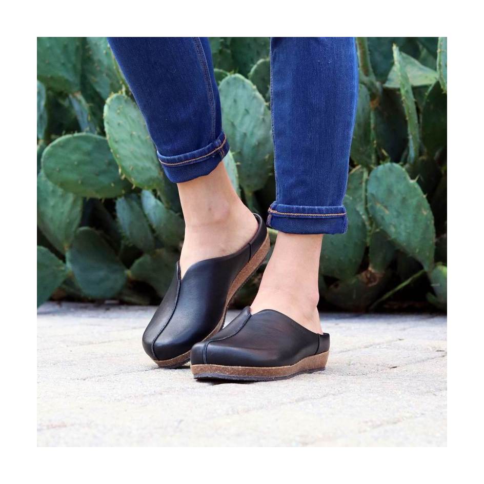 The person is wearing Women's Maria Leather Mule clogs in black, paired with rolled-up blue jeans, standing on a paved surface with cactus leaves in the background.