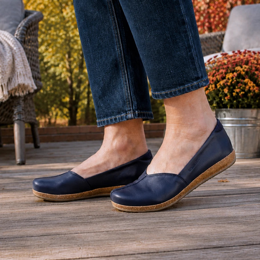 A person wearing the Women’s Liesl Leather Skimmer in dark blue, styled with cuffed jeans, stands on a wooden deck surrounded by autumn plants and outdoor furniture.