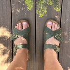 A person in Women's Heidi Leather Sandal with an ultra-cushy footbed stands on a sunlit wooden deck, while tree branches cast shadows above their feet.