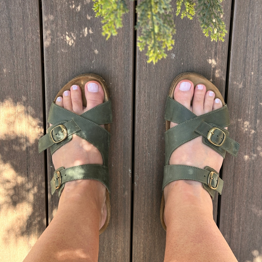 A person in Women's Heidi Leather Sandal with an ultra-cushy footbed stands on a sunlit wooden deck, while tree branches cast shadows above their feet.