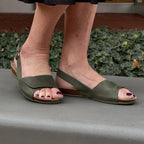 A person wearing Women's Clara Slingback Sandal in olive green, featuring a low wedge heel and cushioned suede footbed, stands outdoors on a gray surface.