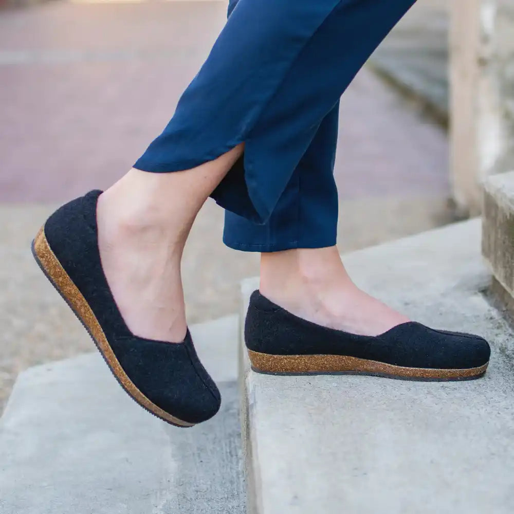 A person wearing navy blue pants and Women's Liesl Wool Skimmer shoes with cork soles and arch support steps up concrete stairs outdoors.
