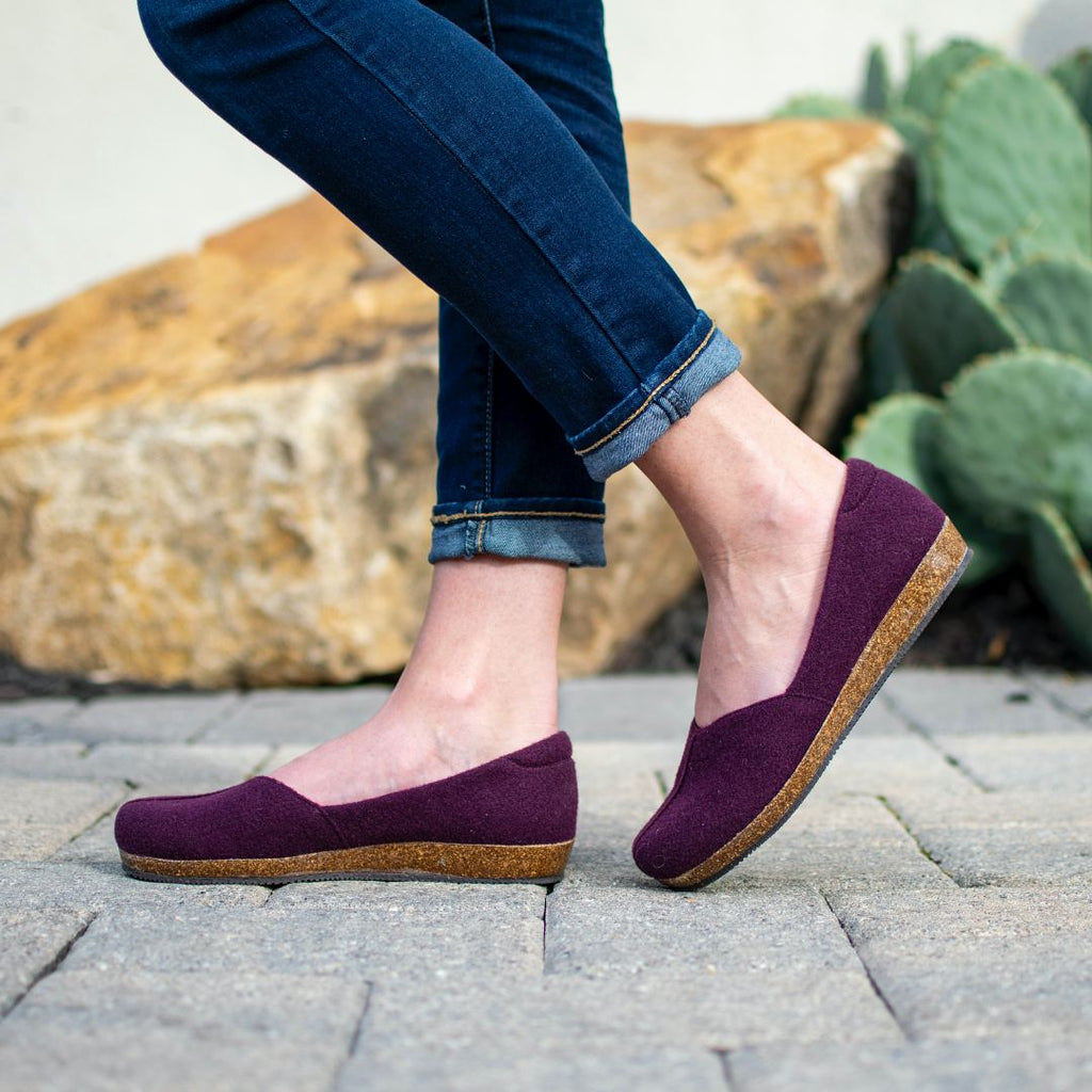 A person in Women's Liesl Wool Skimmer shoes—dark purple wool with cork soles and a wide toe box—paired with rolled-up blue jeans, stands on stone pavement near a cactus and large rocks.