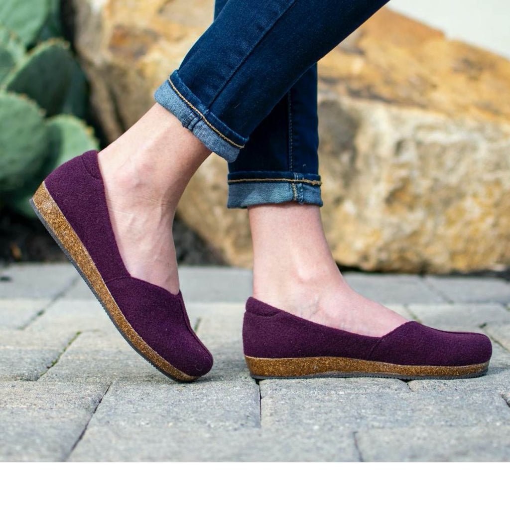 A person wearing Women's Liesl Wool Skimmer in purple, featuring an anatomically shaped support sole, and rolled-up jeans stands on a cobblestone path, surrounded by rocks and cactus.