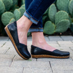 A woman wearing the Women's Liesl Leather Skimmer—black slip-on shoes with a wide toe box and cork soles—paired with cuffed jeans, stands on pavement with prickly pear cacti behind her.