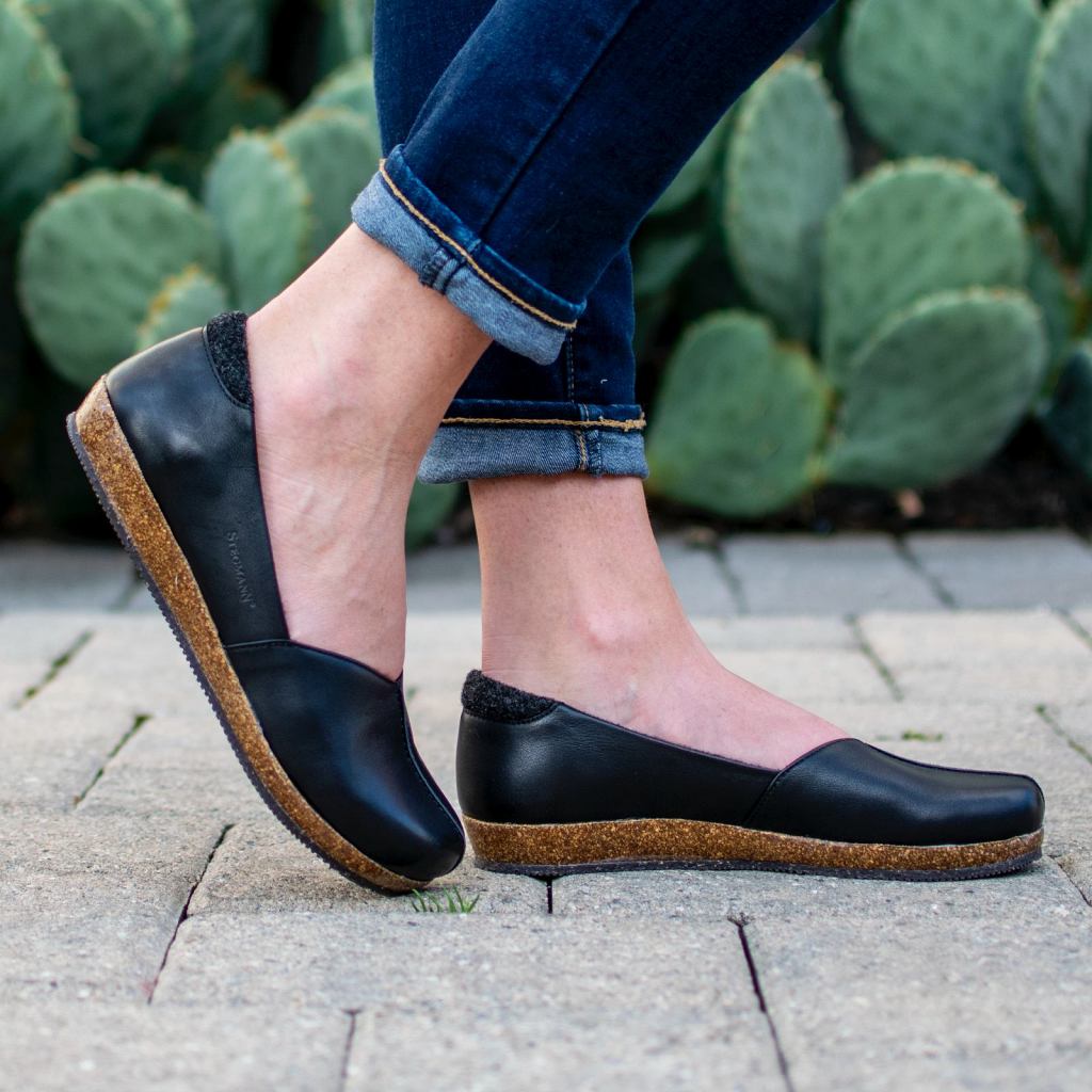 A woman wearing the Women's Liesl Leather Skimmer—black slip-on shoes with a wide toe box and cork soles—paired with cuffed jeans, stands on pavement with prickly pear cacti behind her.